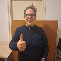 Dorina egy szivárványtortás ajándéktasak előtt guggol, egy lufifújó van a kezében. Feje félrebillentve, szélesen mosolyog. Göndör, barna haját két copfba fogva viseli. Fekete szemüvege, nadrágja és fekete alapon fehér pöttyös pulcsija van. A háttérben türkizkék fitnesslabda.